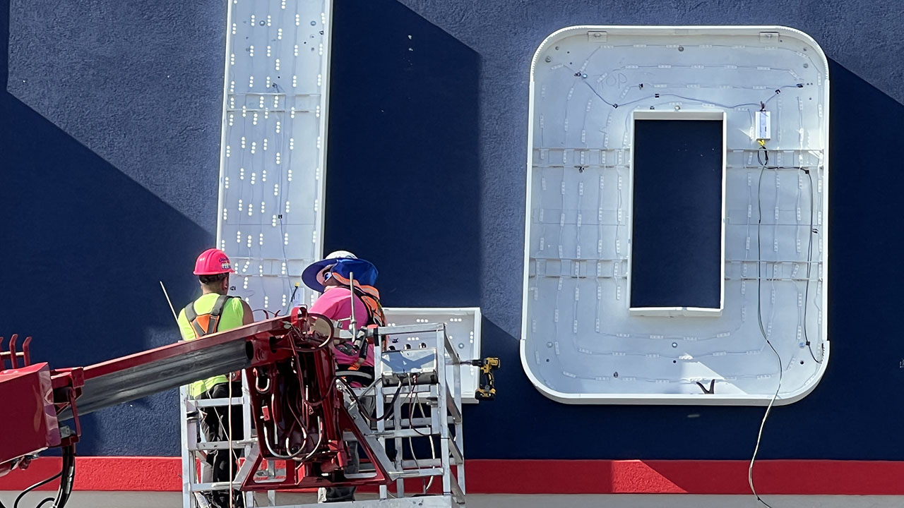 A signage installer stands on a large lift while holding wiring for a GNC Live Well retail sign.