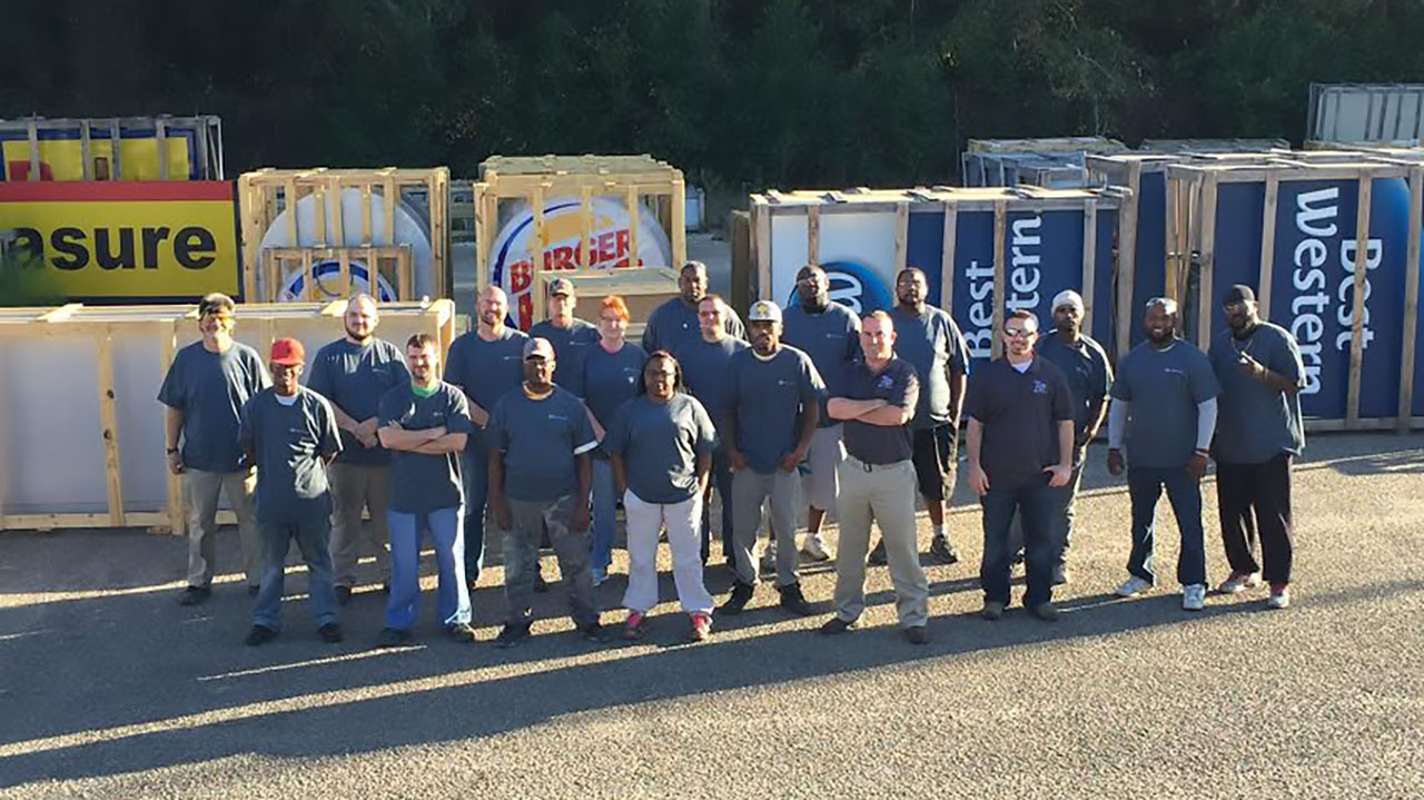 Identiti manufacturing employees posing for a photo outdoors against crates of signs for Burger King, Best Western, and more.