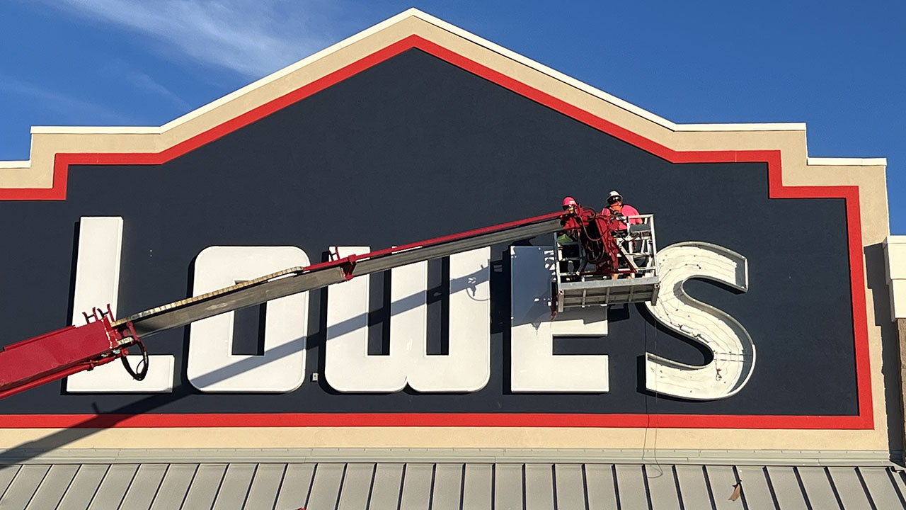 Two workers on a lift operating on a large exterior sign at a Lowe's retail location.