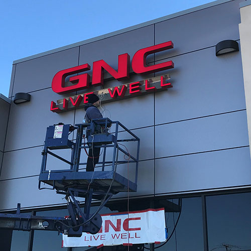 A signage installer stands on a large lift while holding wiring for a GNC Live Well retail sign.